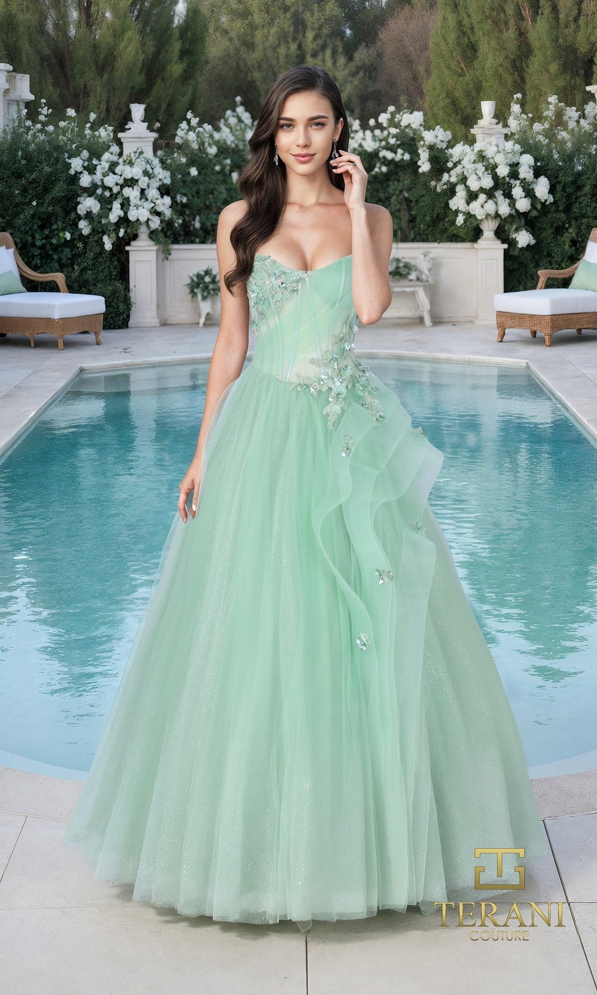 A woman in the celery Terani 261P6197 long formal dress stands by a pool with greenery and outdoor furniture. The strapless gown features sequin lace, floral accents, and layered tulle for an elegant look.